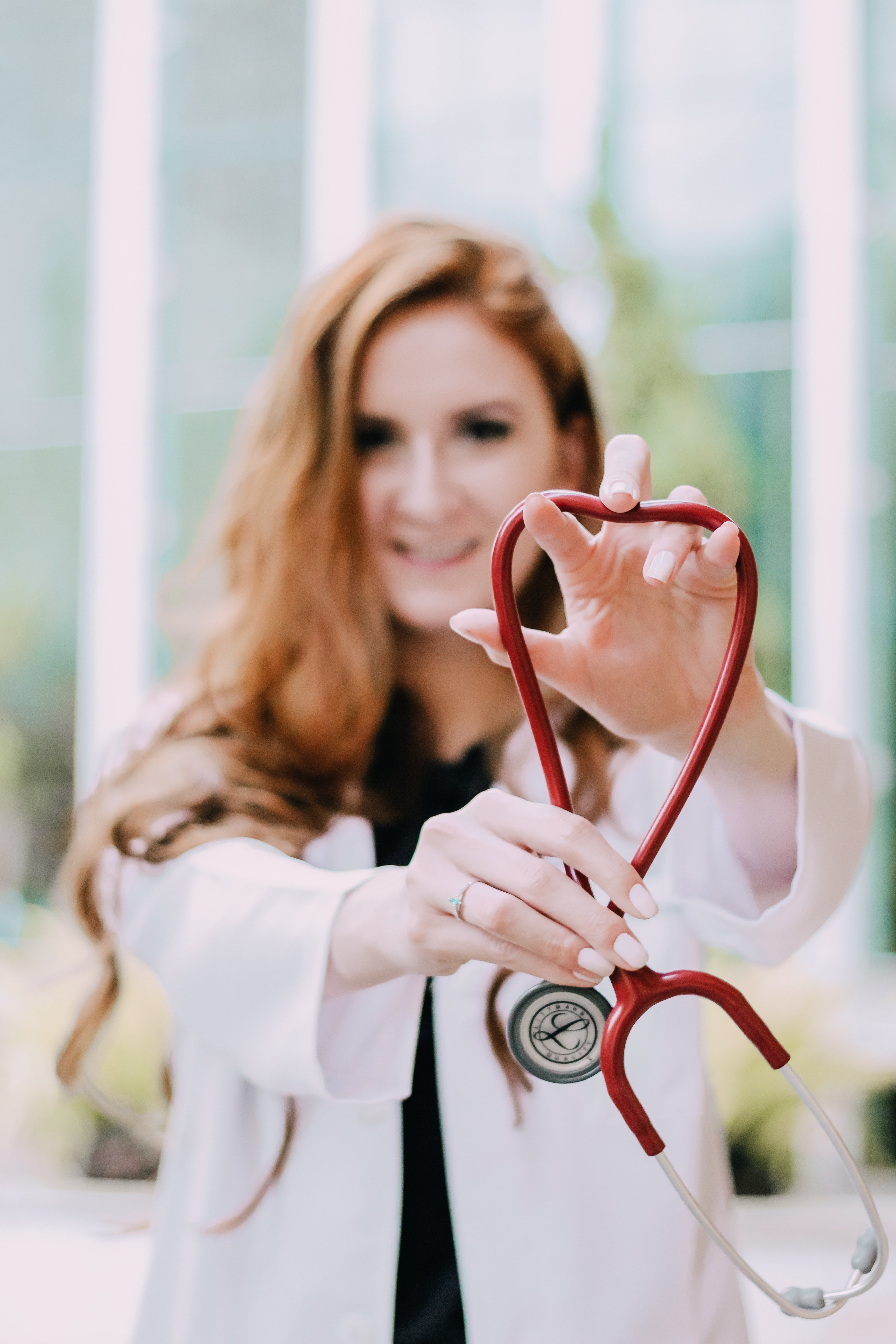 White female doctor holding a red stethoscope towards the camera. The stethoscope hose outlines a heart shape.